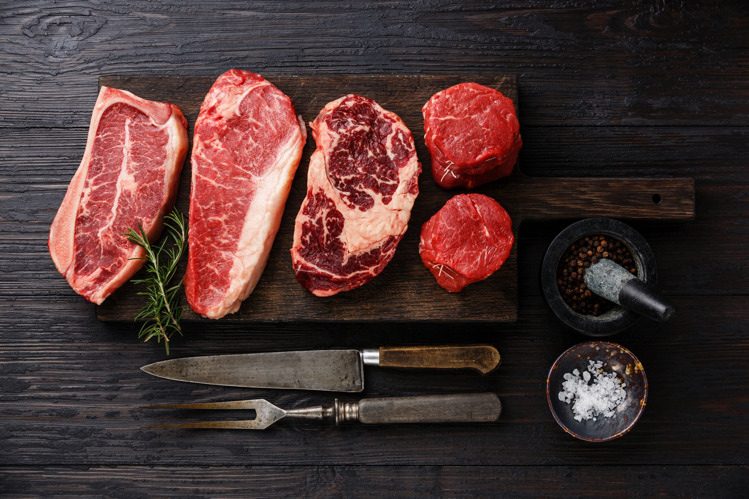 Assorted cuts of raw beef including ribeye, sirloin, and tenderloin arranged on a wooden cutting board, accompanied by a sprig of rosemary, a mortar and pestle with black pepper, and a small bowl of sea salt, with a vintage carving fork and knife.