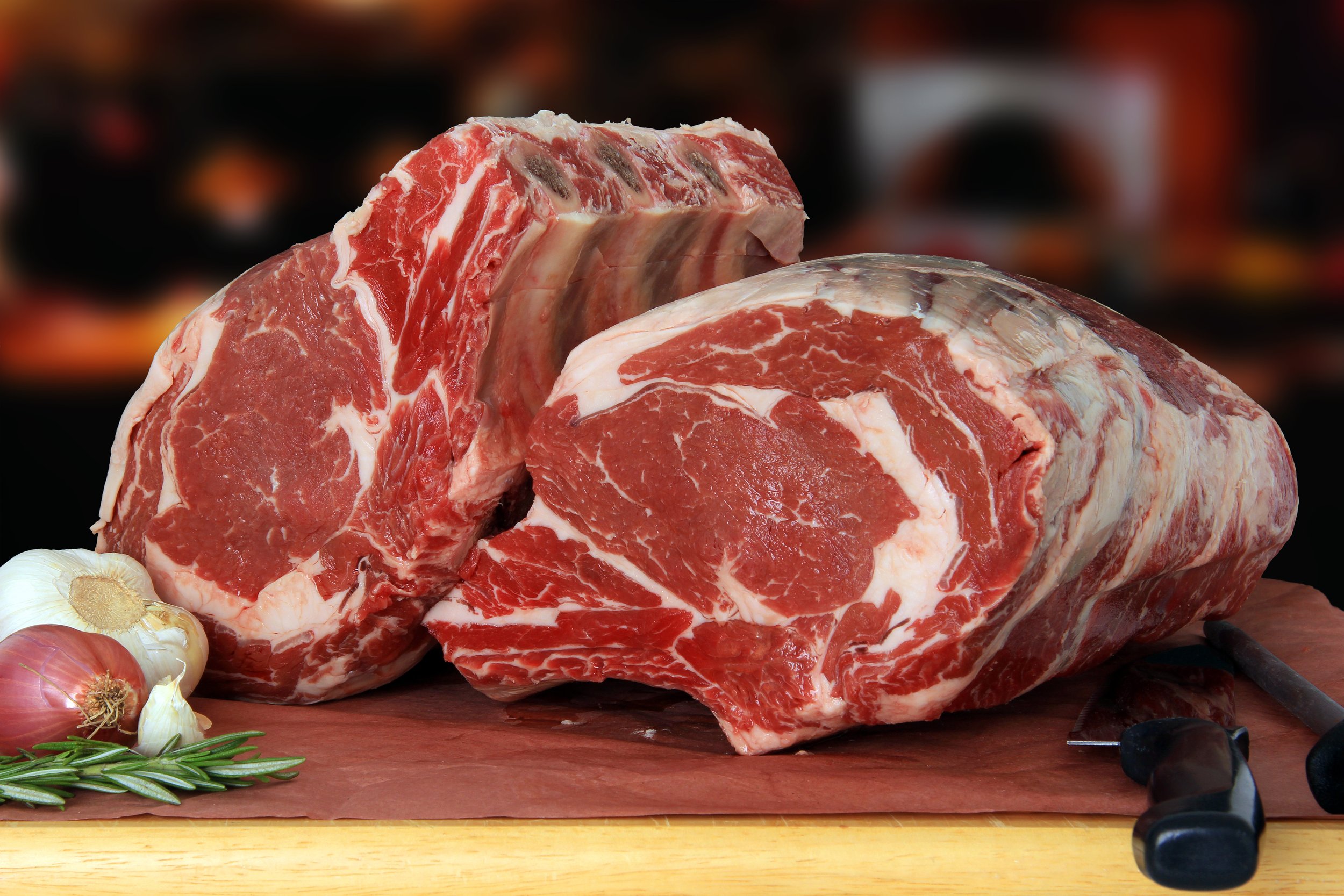 Fresh cuts of ribeye beef with marbling, displayed on butcher paper alongside garlic, shallots, and rosemary, highlighting quality ingredients for gourmet cooking.