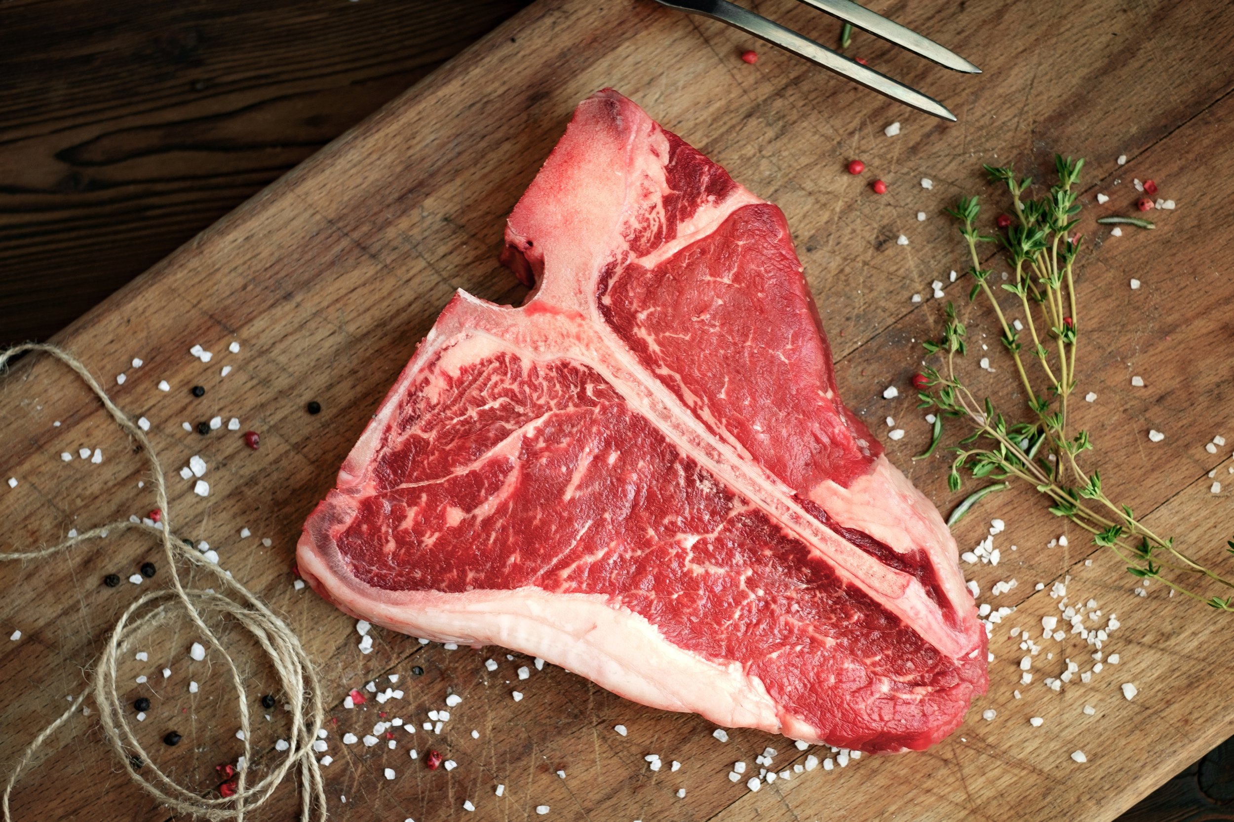 Araw steak resting on a wooden cutting board, surrounded by coarse sea salt, colorful peppercorns, and fresh thyme sprigs, showcasing its marbled texture and rich color.